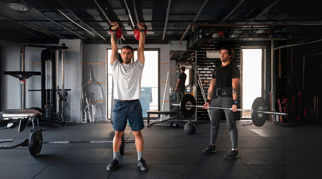 Two men working out in a gym. One man is lifting two kettlebells overhead, while the other is performing a deadlift using a barbell and wearing a black yaghzu lifting belt and wrist straps. 