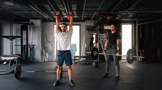 Two men working out in a gym. One man is lifting two kettlebells overhead, while the other is performing a deadlift using a barbell and wearing a black yaghzu lifting belt and wrist straps. 